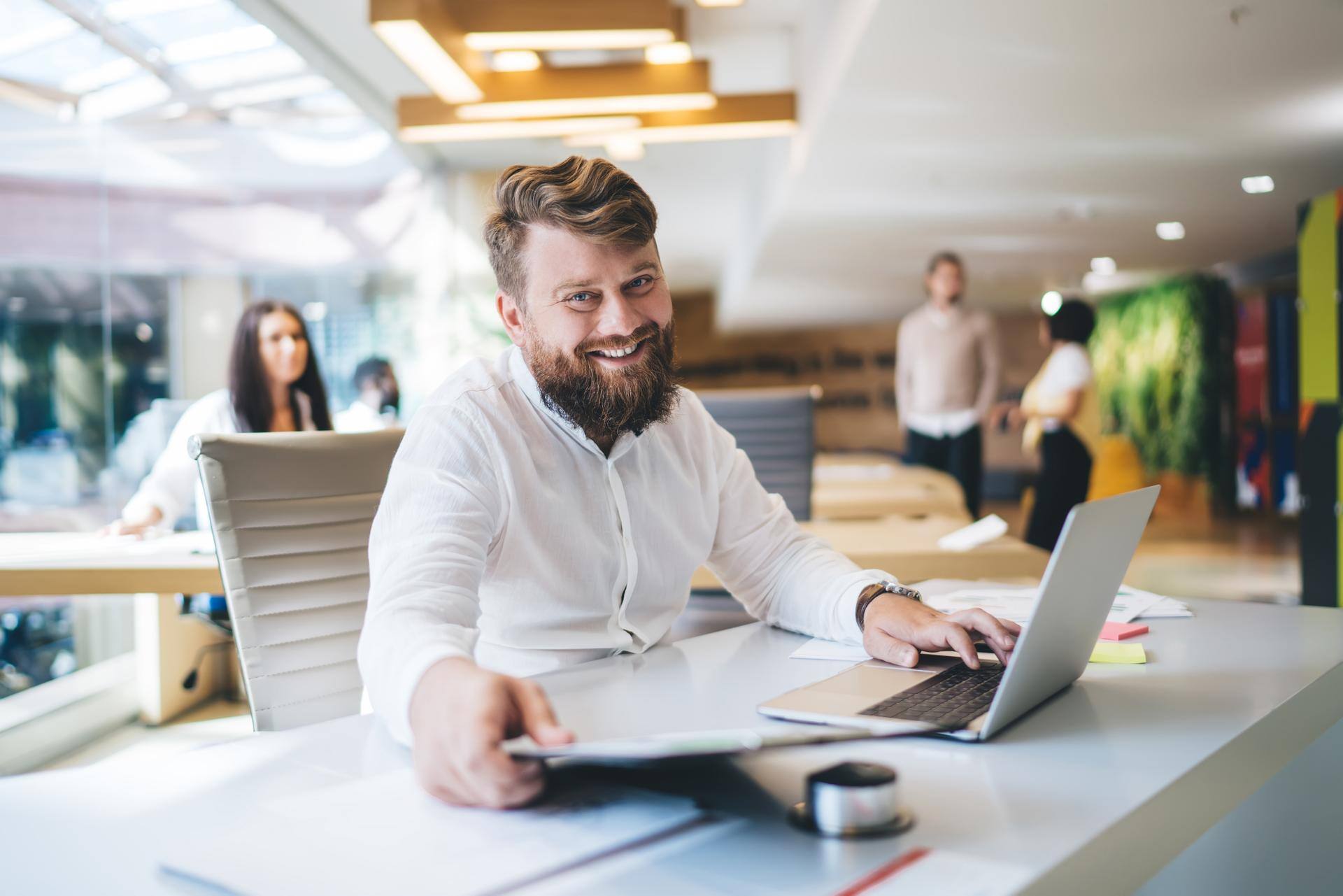 Cheerful businessman in elegant clothing sitting at table in office and using netbook while working on new project and looking at camera